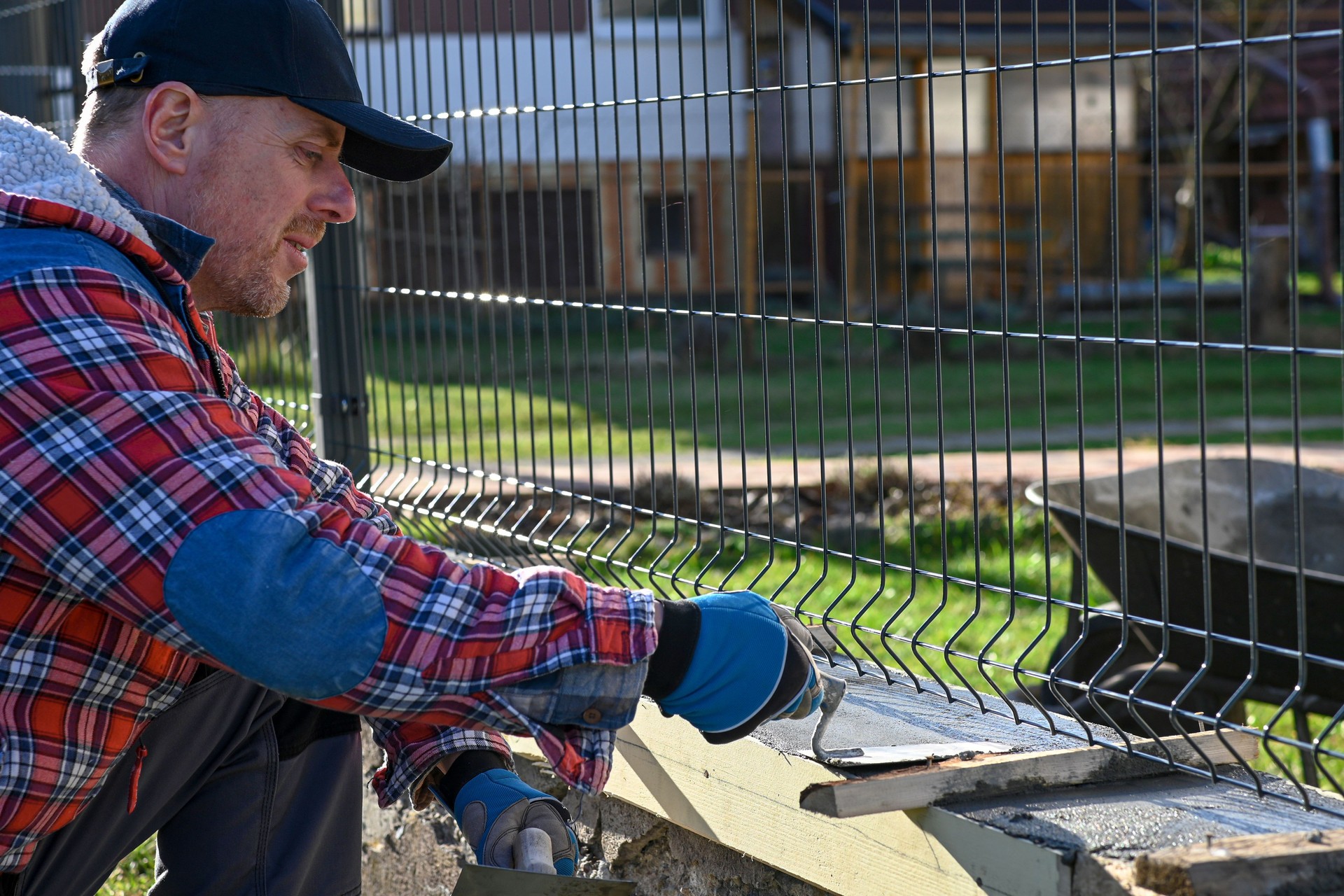 A man in overalls and gloves is repairing the fence in front of the family house. He uses a plane, a trowel and a wheelbarrow.