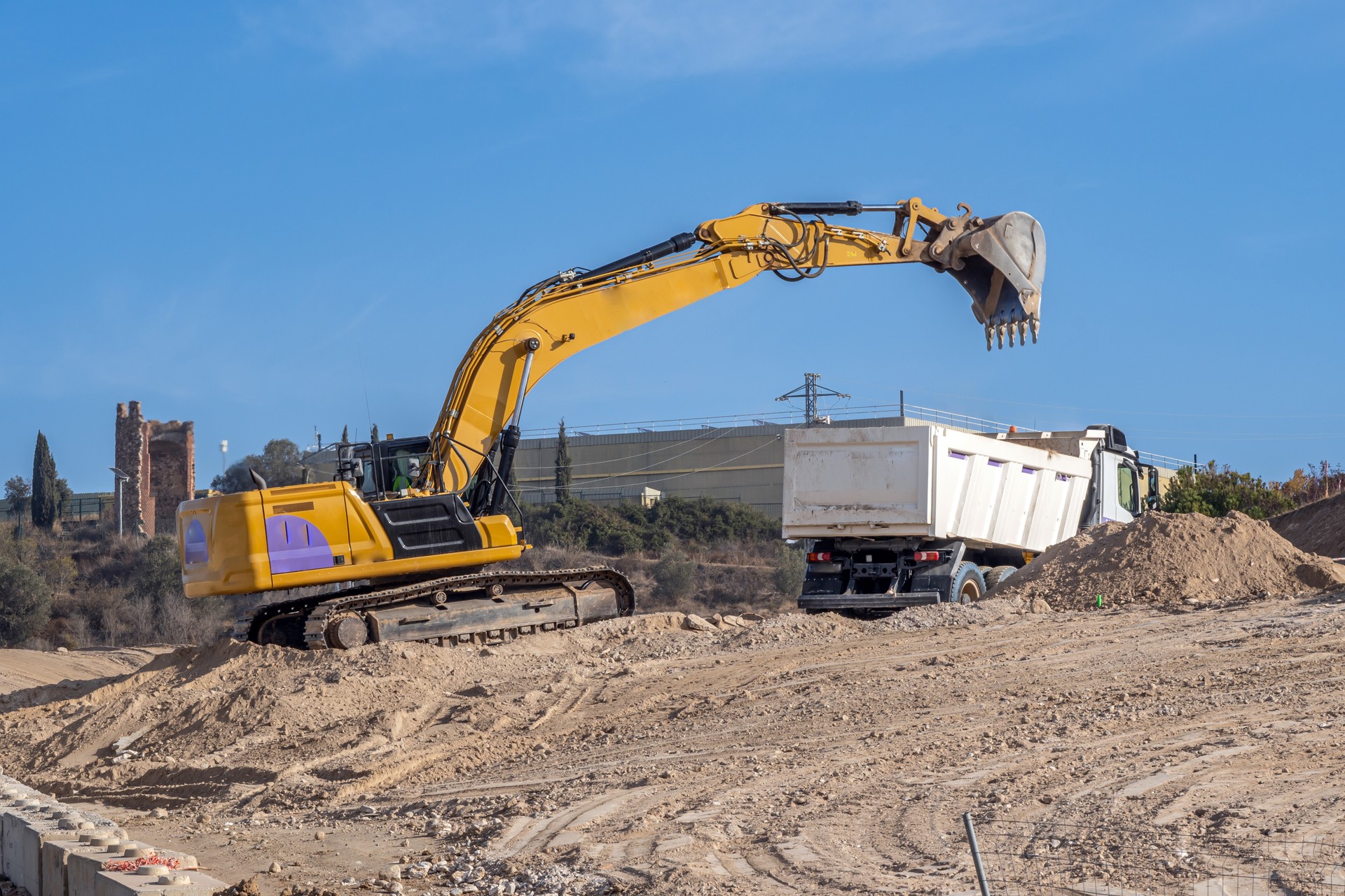 Large yellow excavator depositing sand in a truck bed at a large construction site